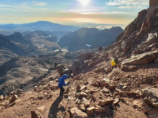 Tourists trekking Mount Kenya through rocky paths on a clear day with vast landscapes behind