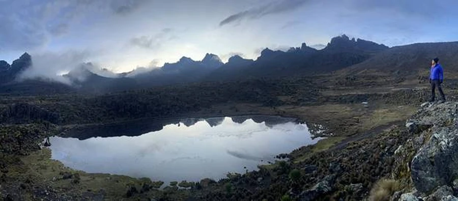 Hiker looking at a Mount Kenya lake with high peaks in the distant as the sun goes down