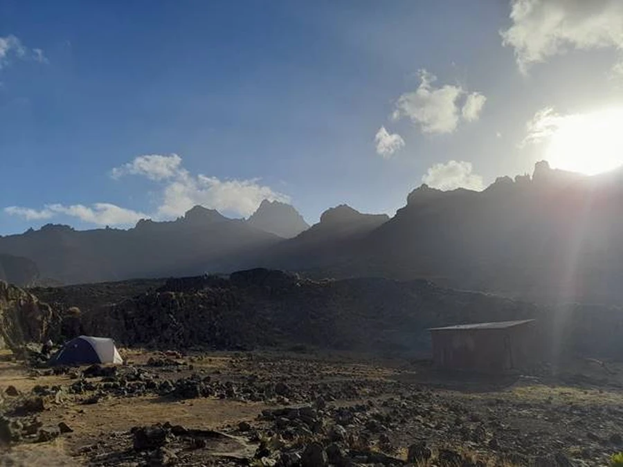 A tent set up close to a cabin on Mount Kenya during a partly cloudy day, peaks in the distant
