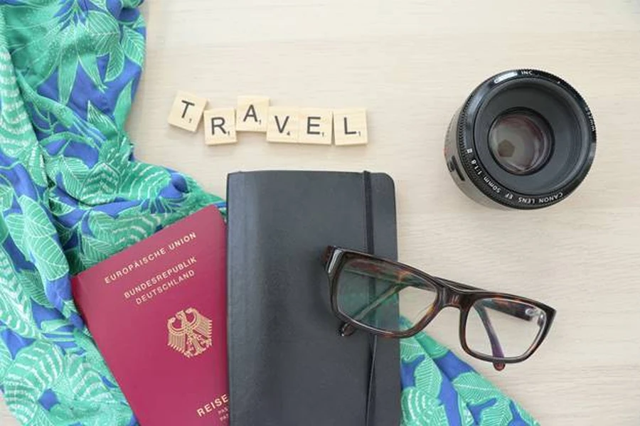 Passport, sunglasses, camera lens, floral fabric, and wallet on a table spread with the word TRAVEL