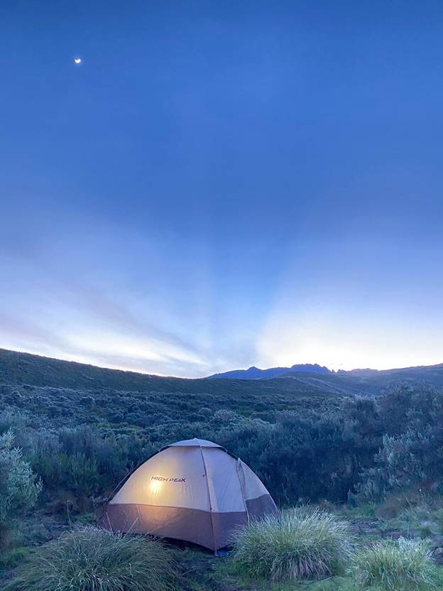 A camp tent with a shining light inside on the slopes of Mount Kenya close to dusk. Peak ahead.