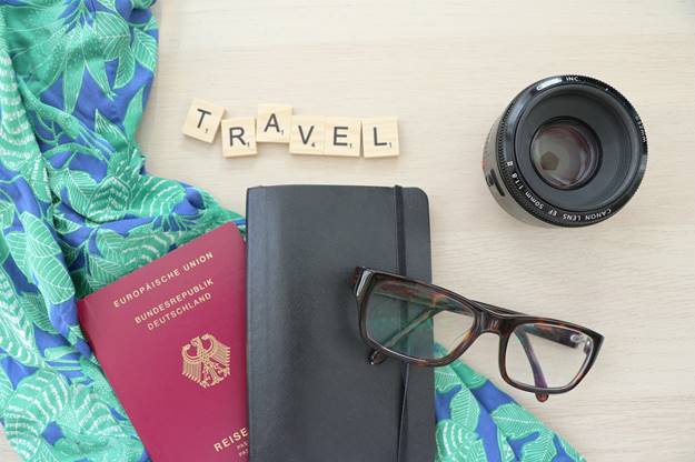 Passport, sunglasses, camera lens, floral fabric, and wallet on a table spread with the word TRAVEL