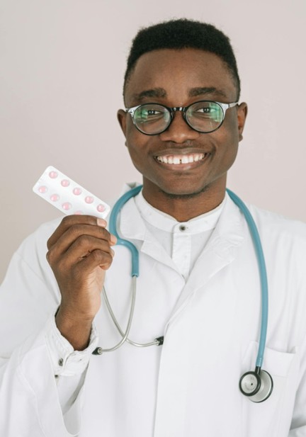 A doctor in white lab coat and stethoscope smiling and holding a medication packet