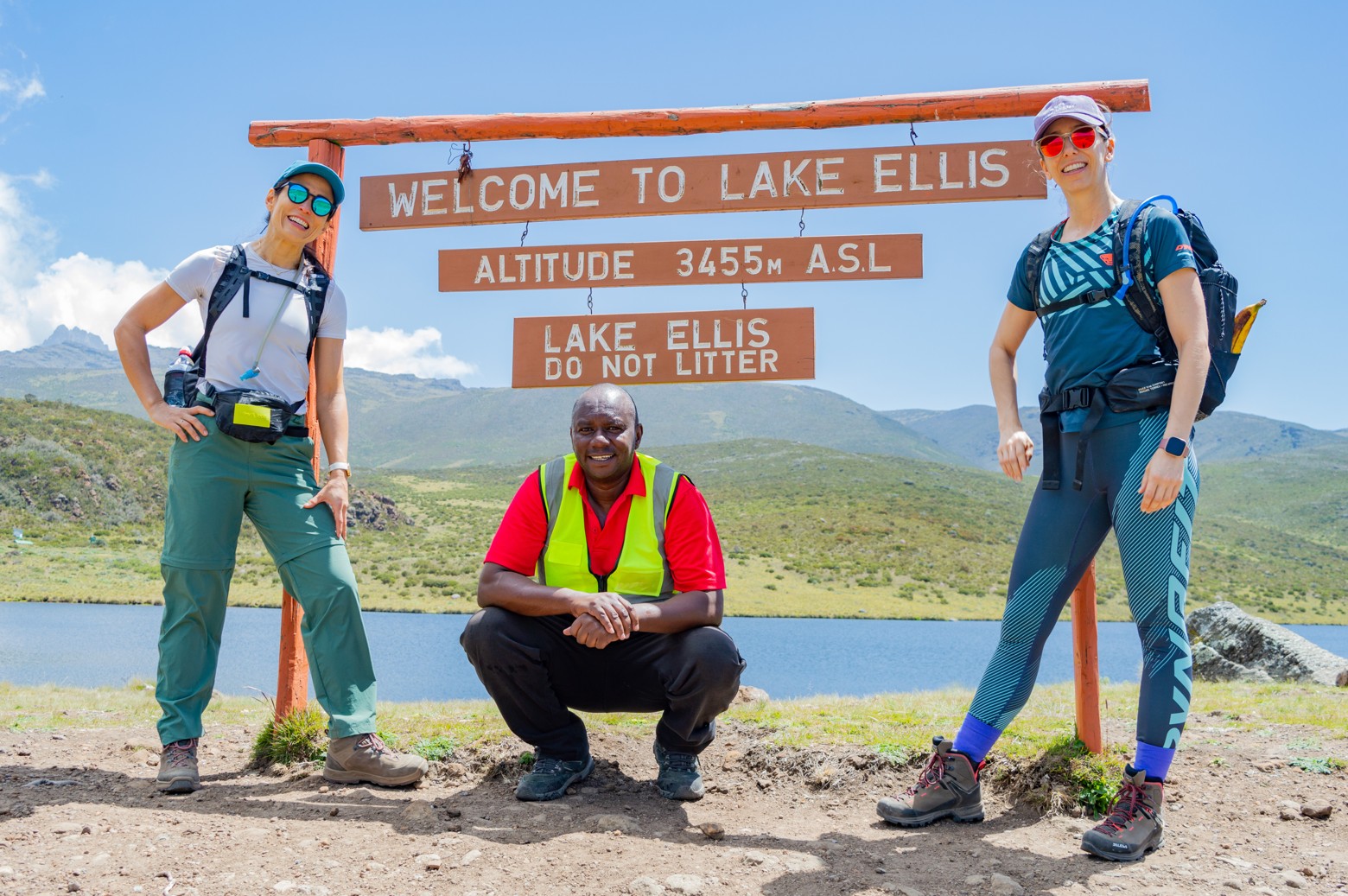 Two hikers and a guide at the Lake Ellis signpost on Mount Kenya with the lake behind them